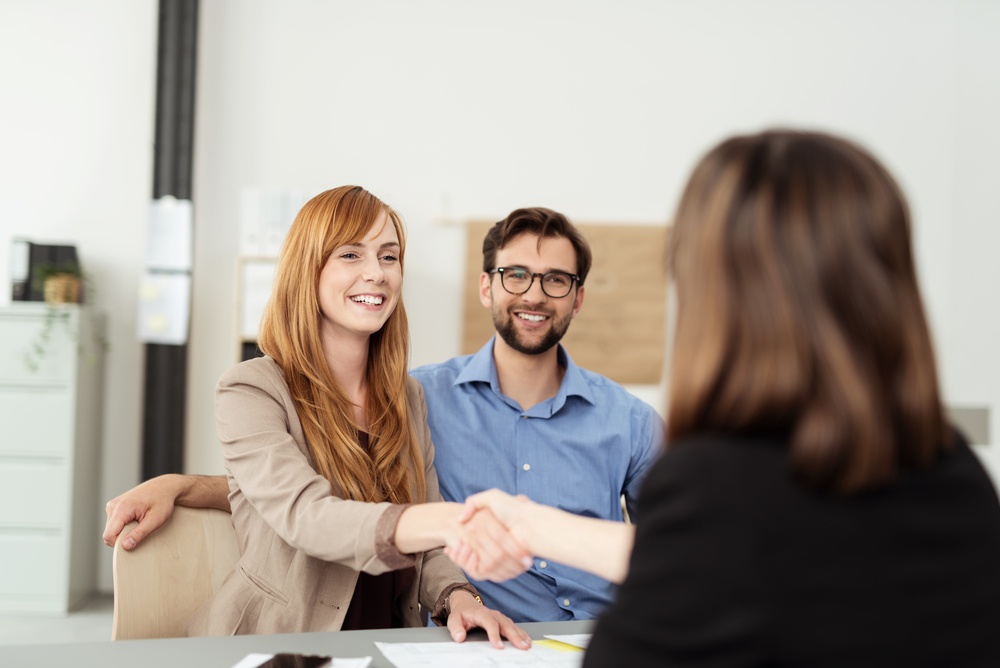 Happy young couple meeting with a broker in her office leaning over the desk to shake hands, view from behind the female agent.jpeg